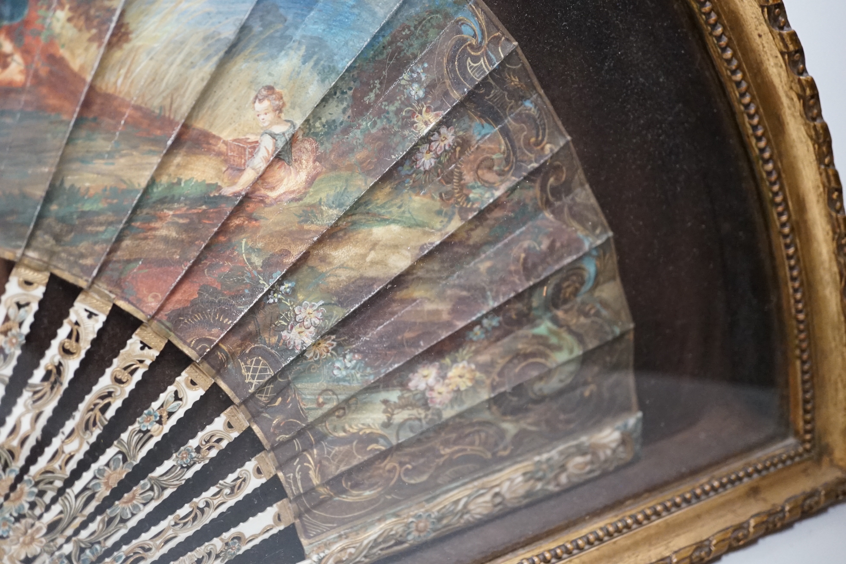 A 19th century French bone fan with pierced decoration, hand painted with figures wearing 18th century dress before a landscape, framed, the frame 62cm wide